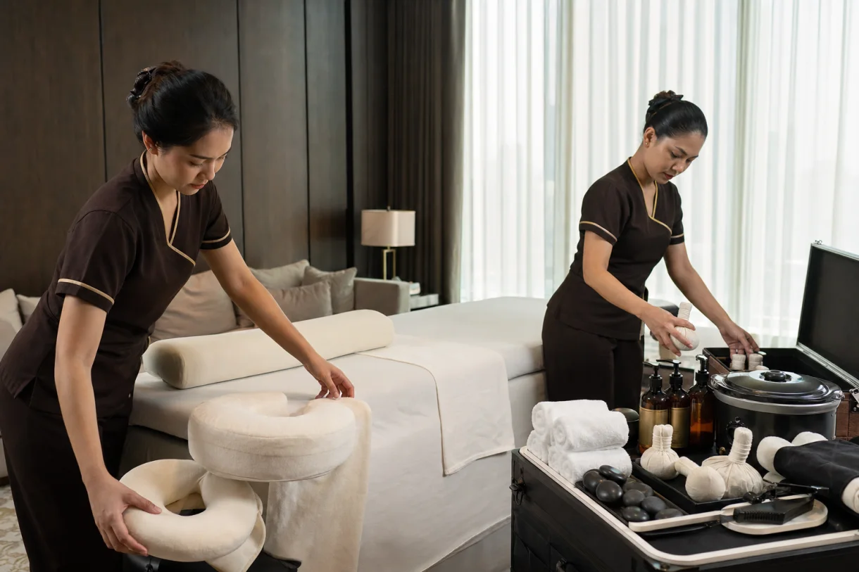 Two therapists arranging linens, hot stones, herbal compresses and professional equipment for a Bangkok home spa
